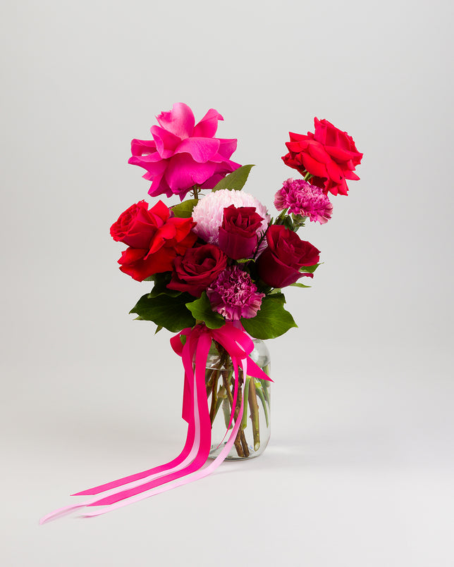 A bouquet of red and pink roses with some additional flowers in a vase, with decorative ribbons.