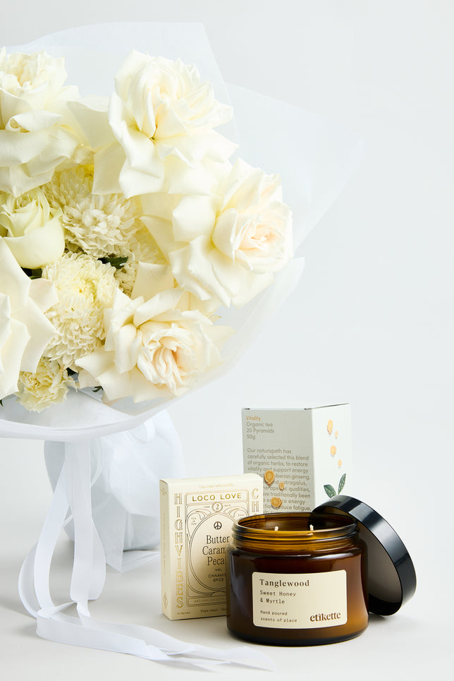 Pure Grace Gift Set by Flowerdose — a timeless white flower bouquet featuring premium ivory roses, white chrysanthemums, and soft-toned blooms, elegantly wrapped in white paper with satin ribbons. Paired with a Tanglewood Sweet Honey & Myrtle candle, Loco Love Butter Caramel Pecan chocolate, and Vitality organic tea — a luxury Melbourne florist gift perfect for birthdays, thank-you gestures, and elegant occasions.