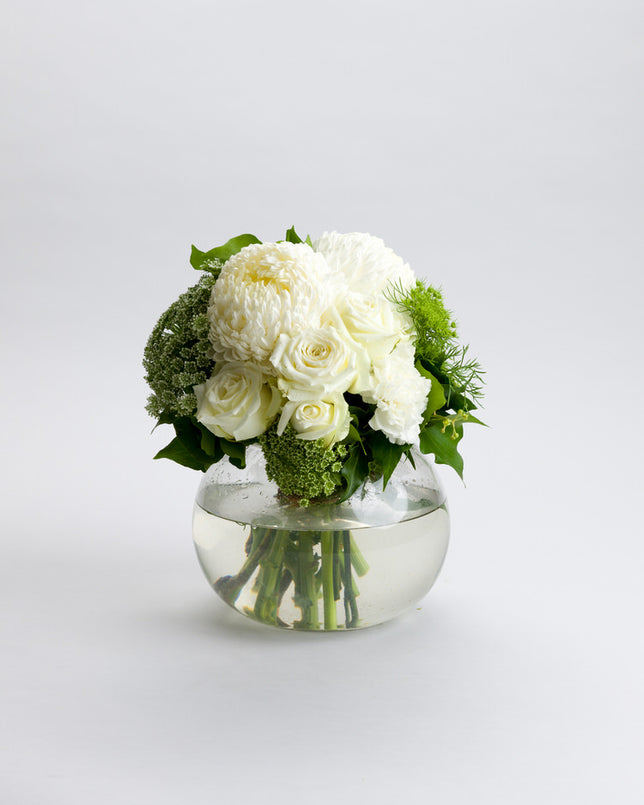 A white flower arrangement including roses and other foliage, presented in a clear glass fish-bowl vase.